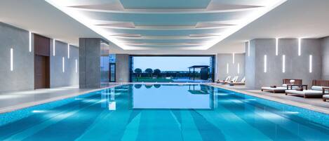 En moderne indendørs swimmingpool med krystalklart udsyn til himlen og et loungeområde ved poolen.