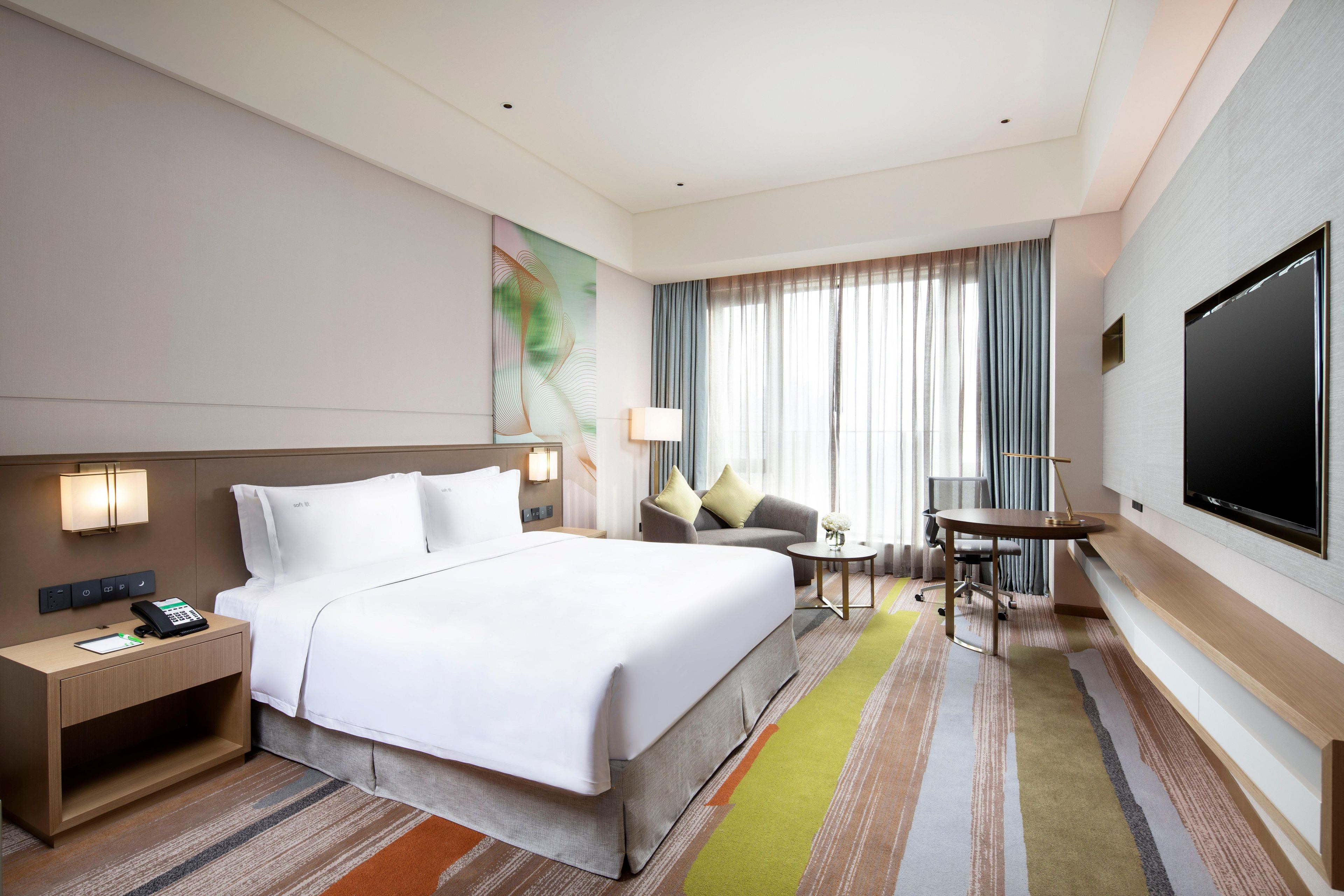 A hotel room with a large bed, a desk, a chair, a TV, and a window with curtains.