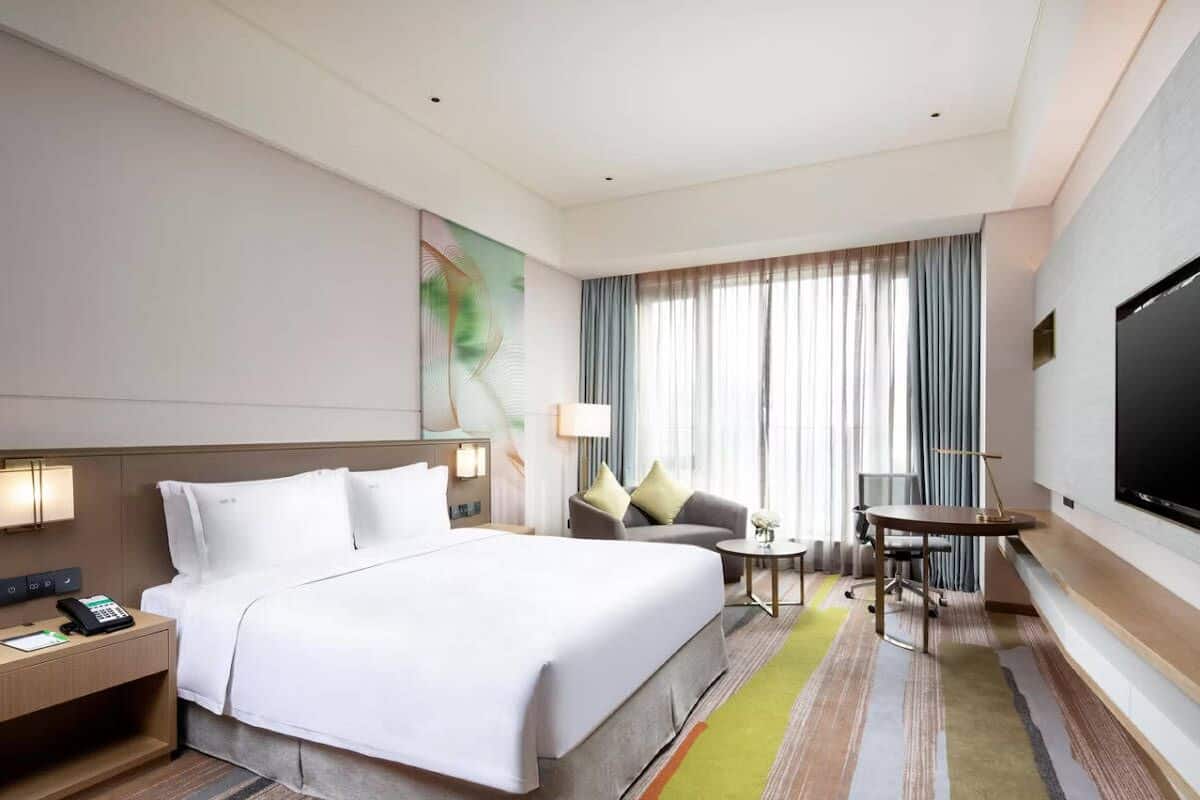 A hotel room with a large bed, a desk with a telephone, a small table with a lamp, a television, and a window with curtains.