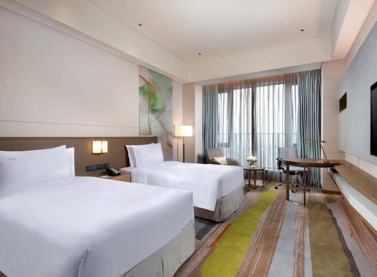 A hotel room with two beds, a desk, a chair, a lamp, and a large window with curtains.
