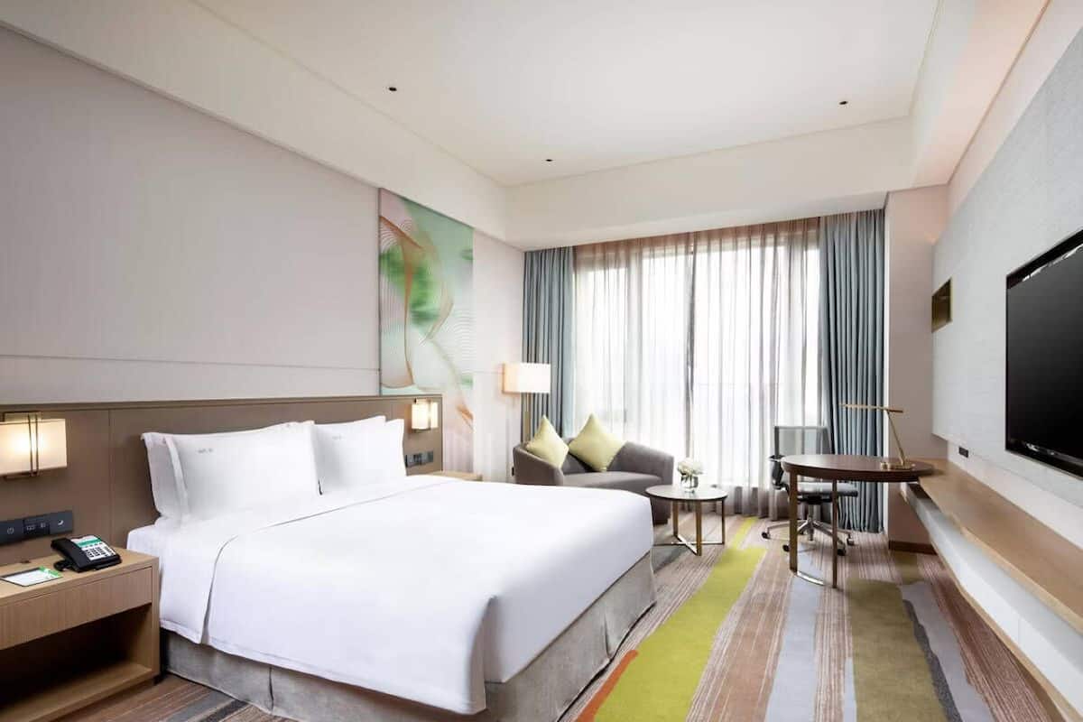 A hotel room with a large bed, a desk with a telephone, a small table with a lamp, and a TV on the wall.