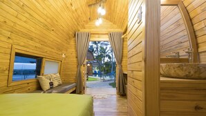 A wooden cabin room with a bed, a window, a sink, and a view of a pool and palm trees. - CoCo Farm Hoi An (Da Nang)