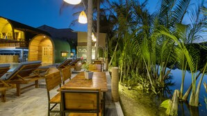 A wooden outdoor seating area with lounge chairs and tables, surrounded by palm trees and a body of water. - CoCo Farm Hoi An (Da Nang)