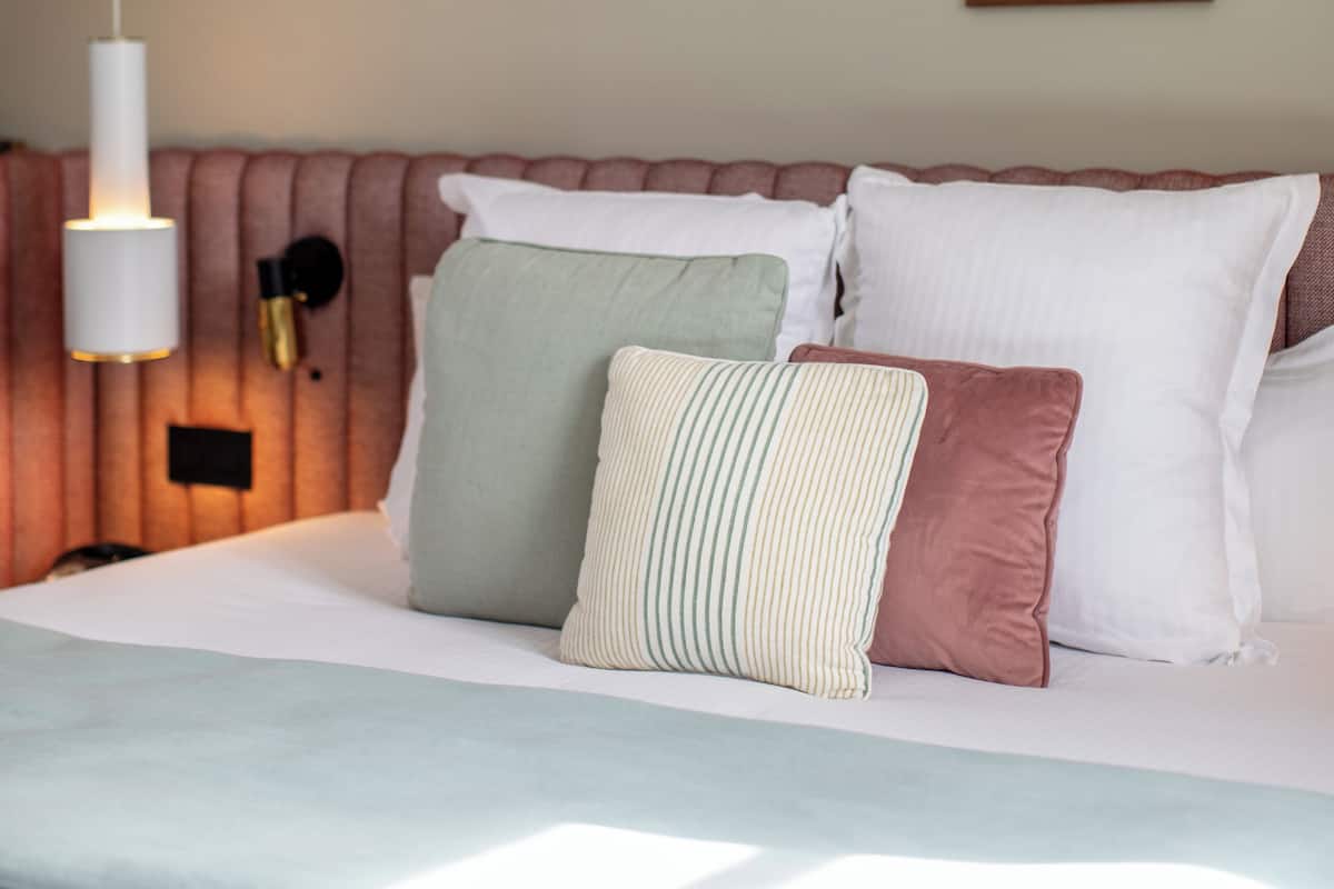 A neatly made bed with a striped pillow, a pink pillow, and a green pillow. There is a framed picture of a person in water on the wall.