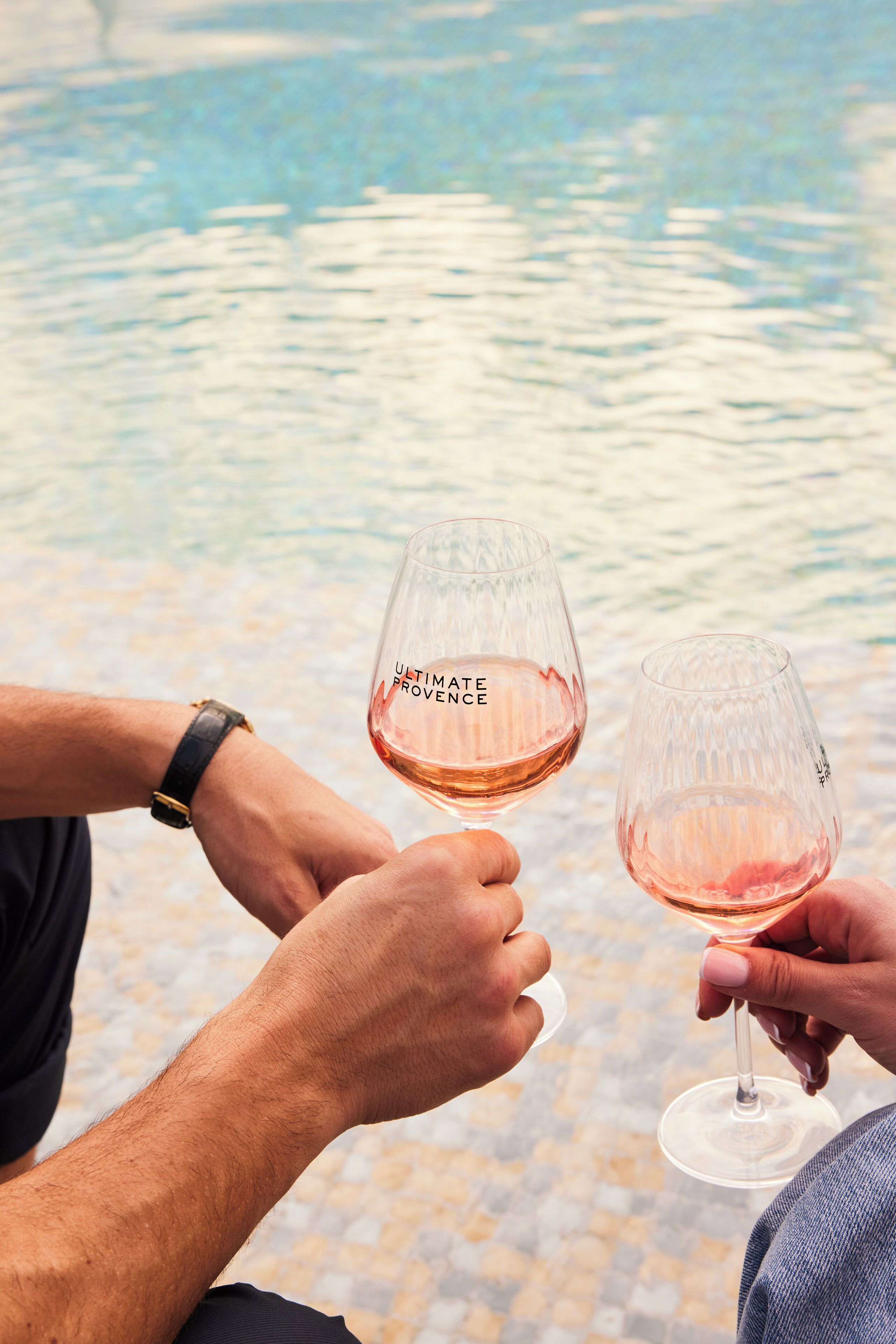 Two people holding wine glasses with the text "ULTIMATE PROVENCE" on them, standing by a body of water.