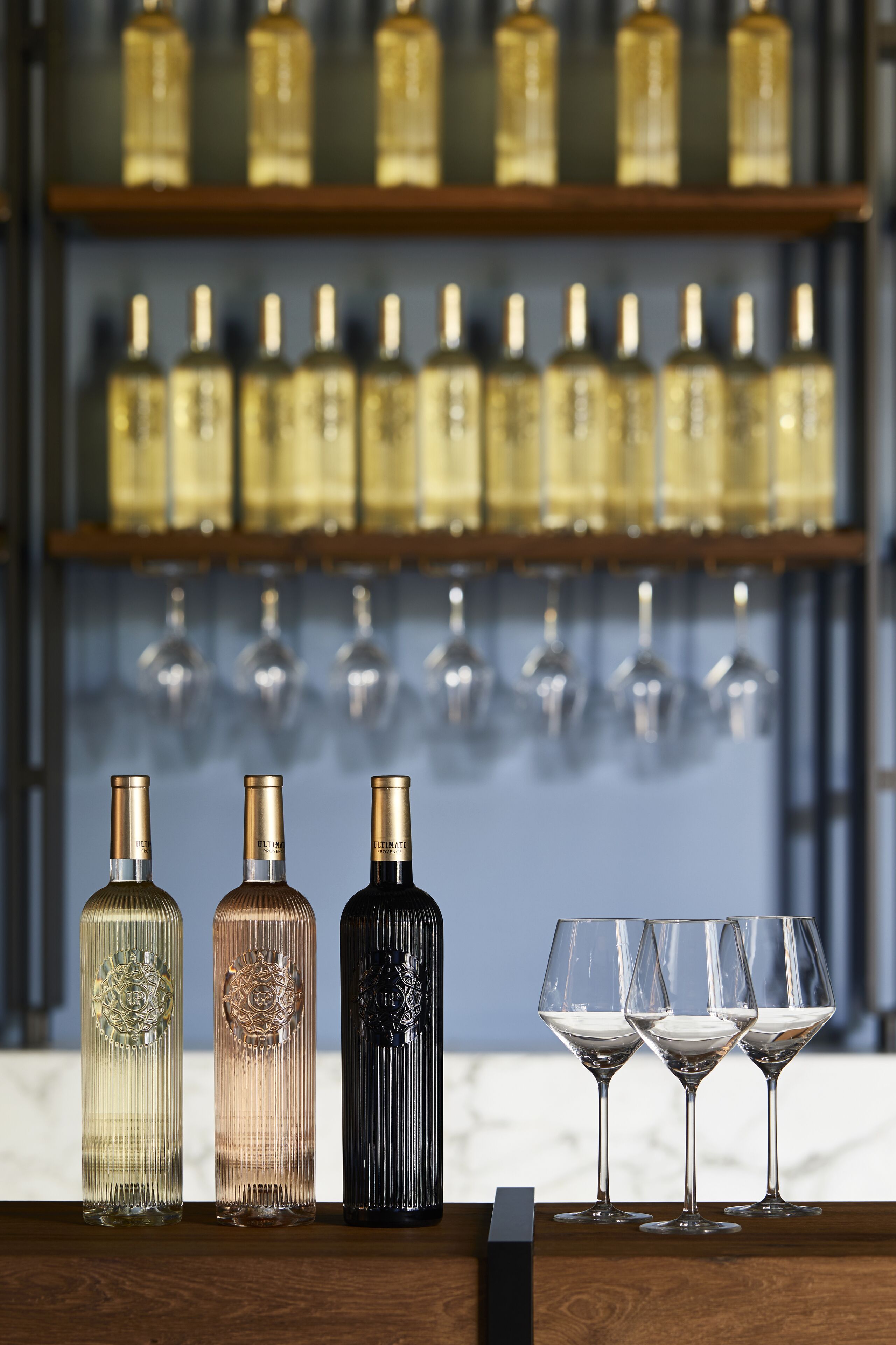 Three bottles and three glasses on a wooden surface with a shelf of bottles and glasses in the background.