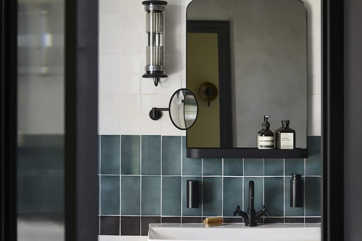 A modern bathroom with a white sink, black faucet, and a mirror above it. There are two bottles on the mirror's shelf and towels hanging below the sink.