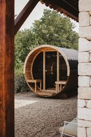 Una estructura cilíndrica de madera con un banco en su interior, ubicada frente a un muro de piedra y rodeada de grava.