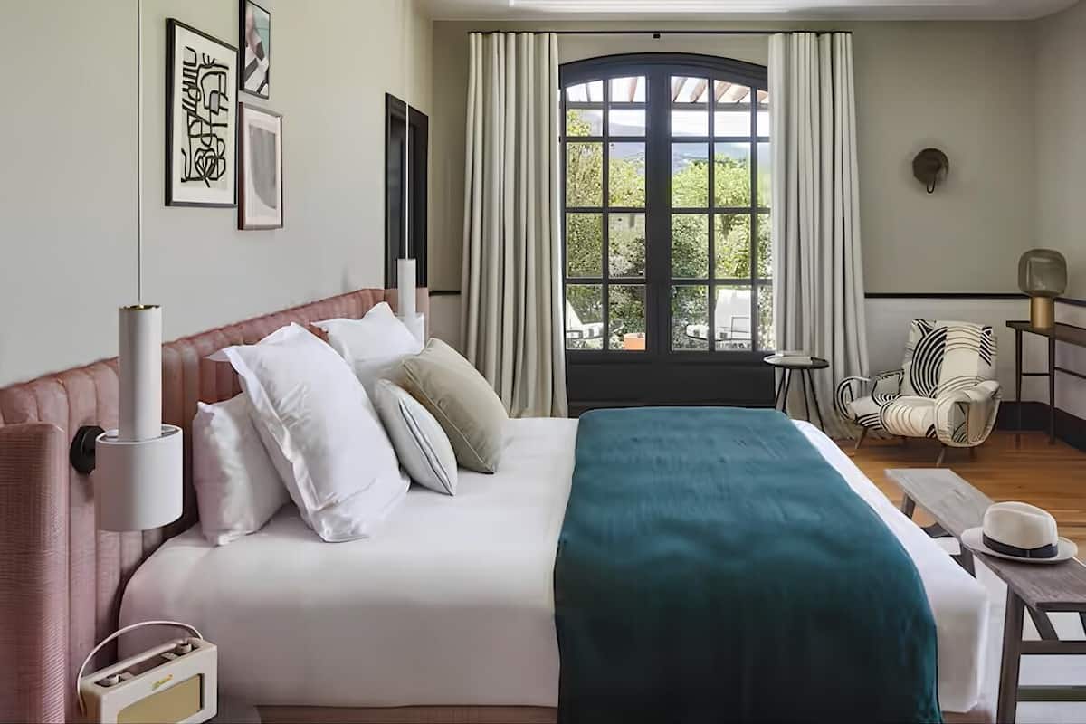 A hotel room with a bed, a window with curtains, a chair, and a lamp.