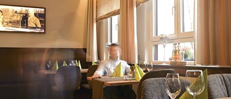 Interior de un restaurante con una mesa para dos, un televisor en la pared y ventanas amplias con cortinas.