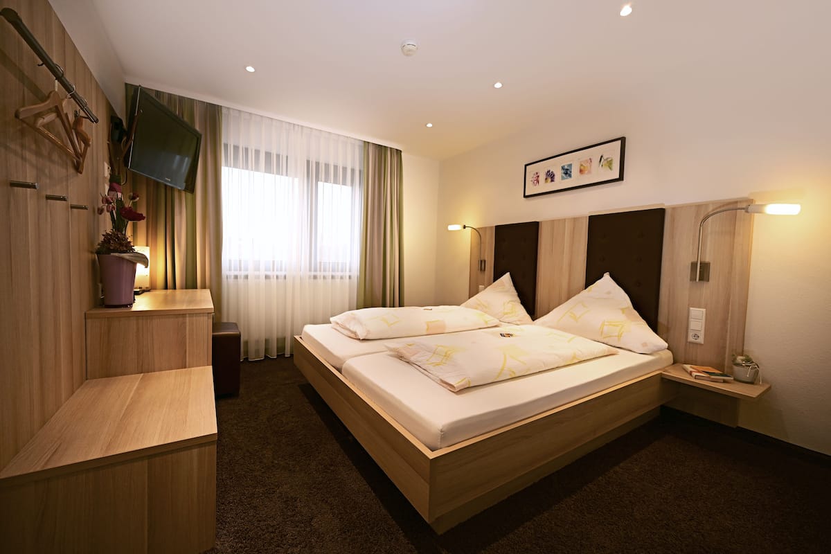 A modern hotel room with a large bed, a TV mounted on the wall, a bedside table with a lamp, and a window with sheer curtains.
