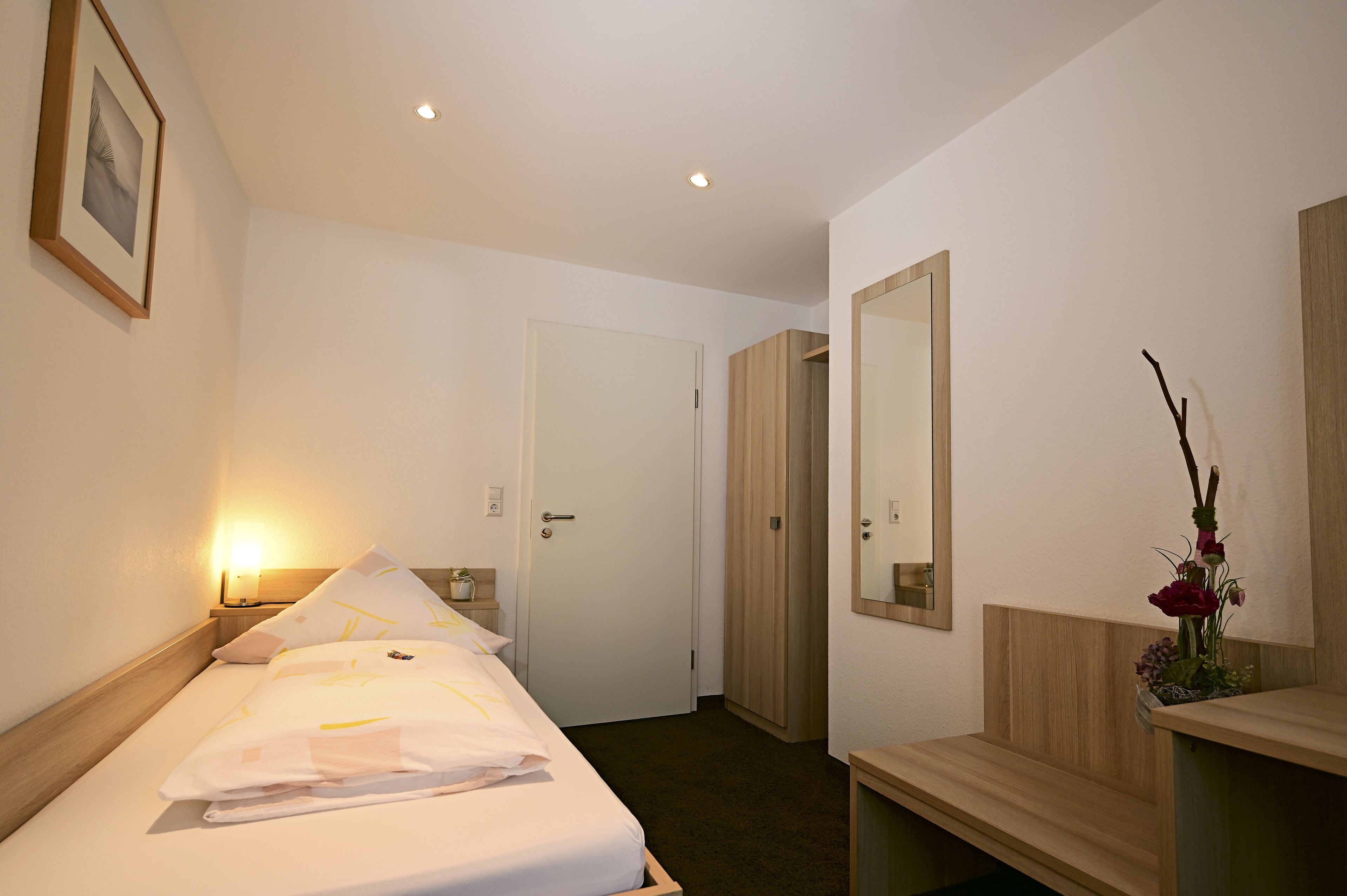 Ein Hotelzimmer mit Bett, Nachttisch, Lampe, Spiegel und einer kleinen Pflanze.