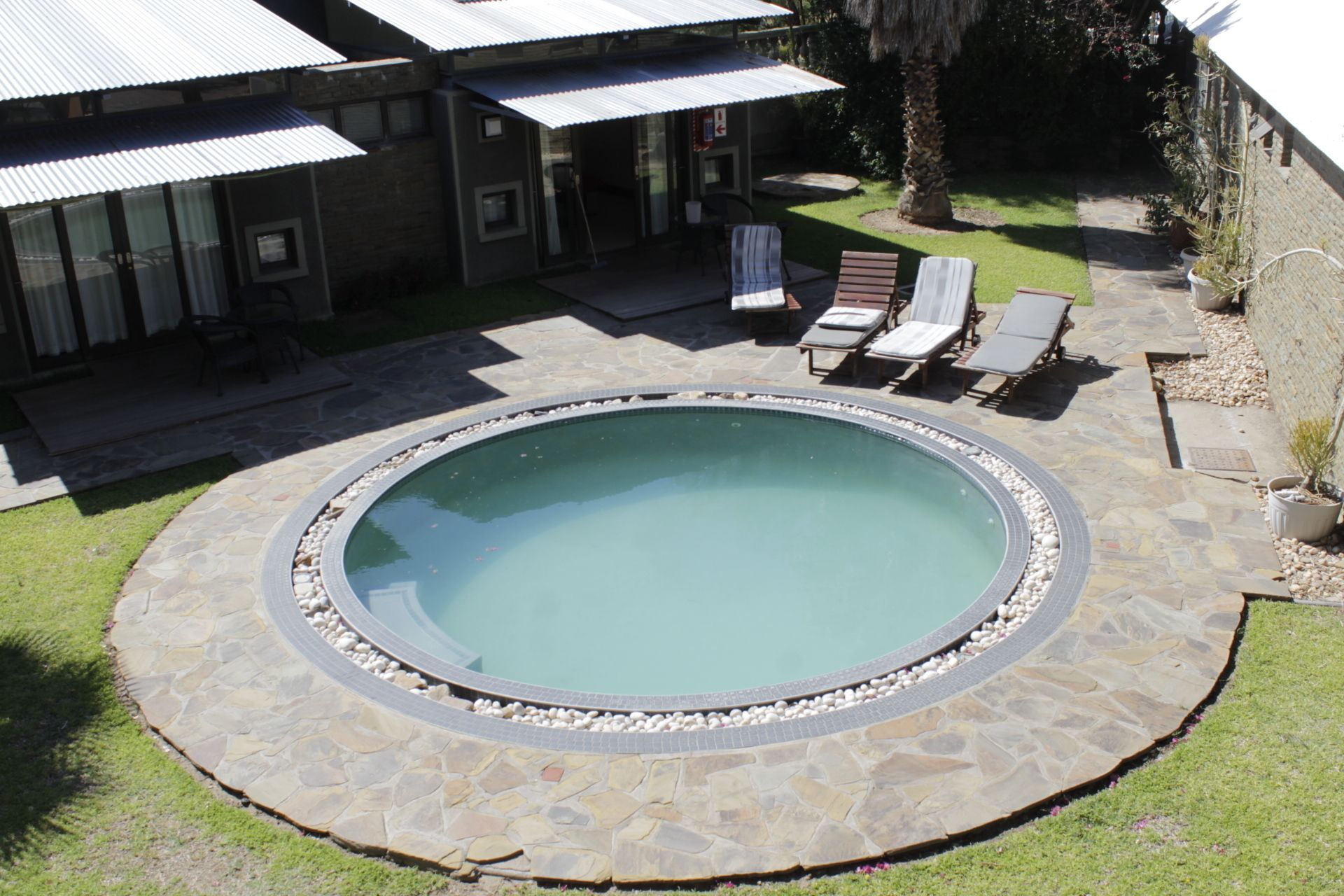 Una piscina circular con borde de piedra, rodeada por una zona de patio con sillones de descanso y un pequeño jardín.
