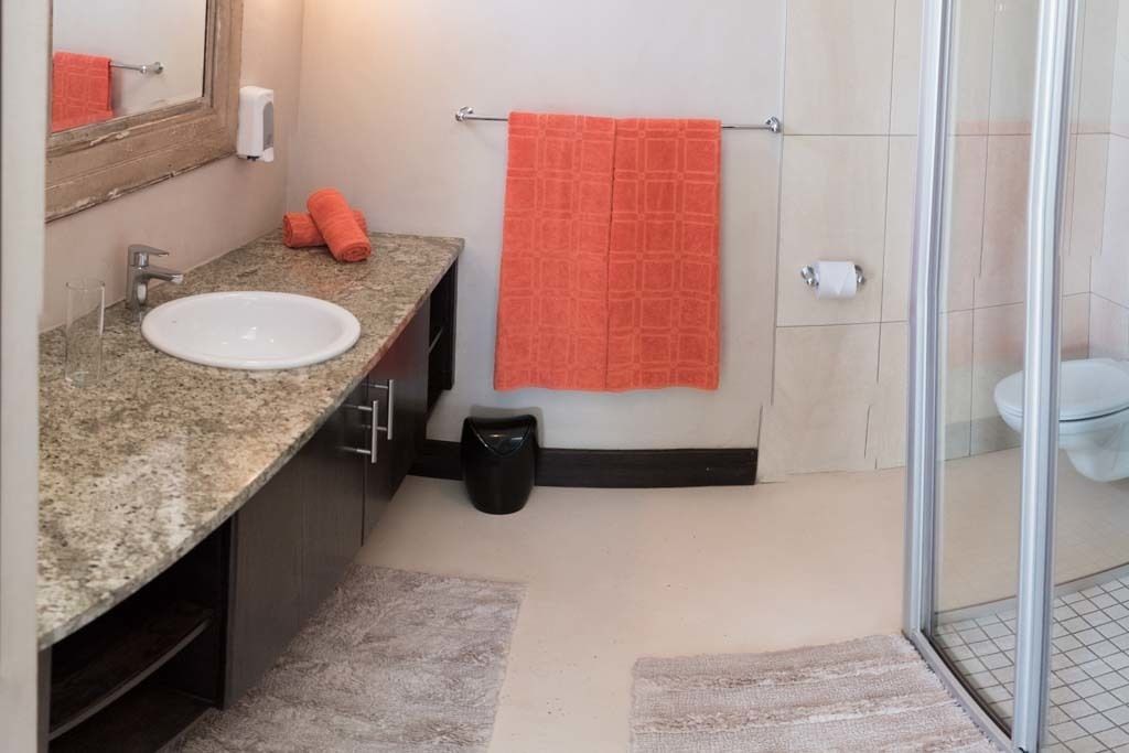 A bathroom with a granite countertop sink, a mirror, a towel rack with an orange towel, a trash bin, and a glass shower enclosure.