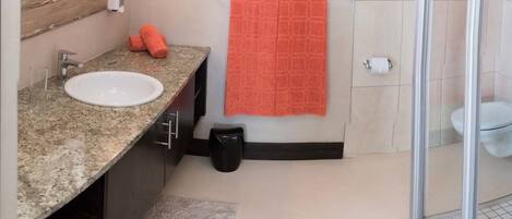 A bathroom with a granite countertop sink, a mirror, a towel rack with an orange towel, a trash bin, and a glass shower enclosure.