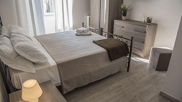 A neatly made bed with a folded towel, a bedside lamp, and a dresser with a plant. - Grandis Rooms (Rome)