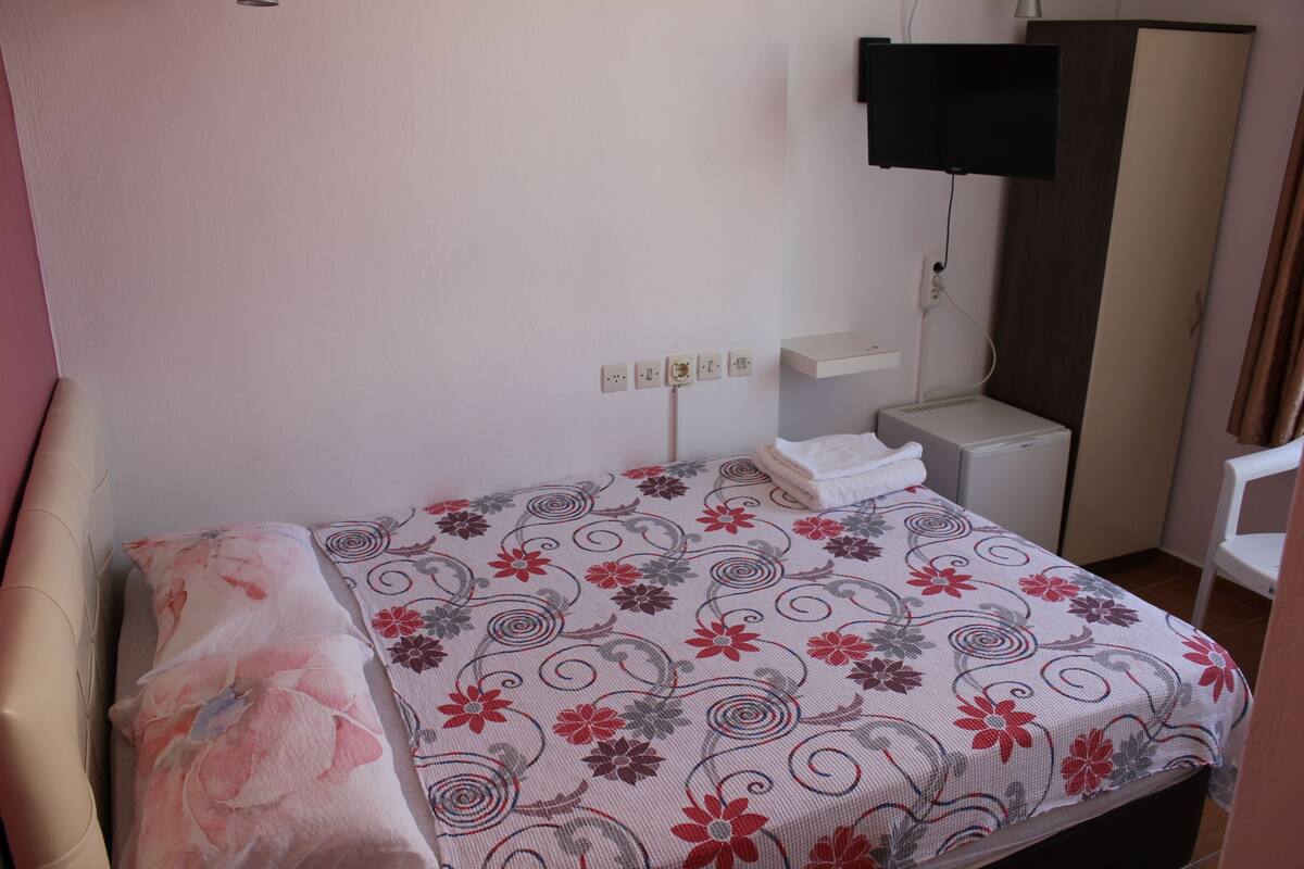 A bedroom with a bed, a TV mounted on the wall, a wardrobe, and a small table with a lamp.