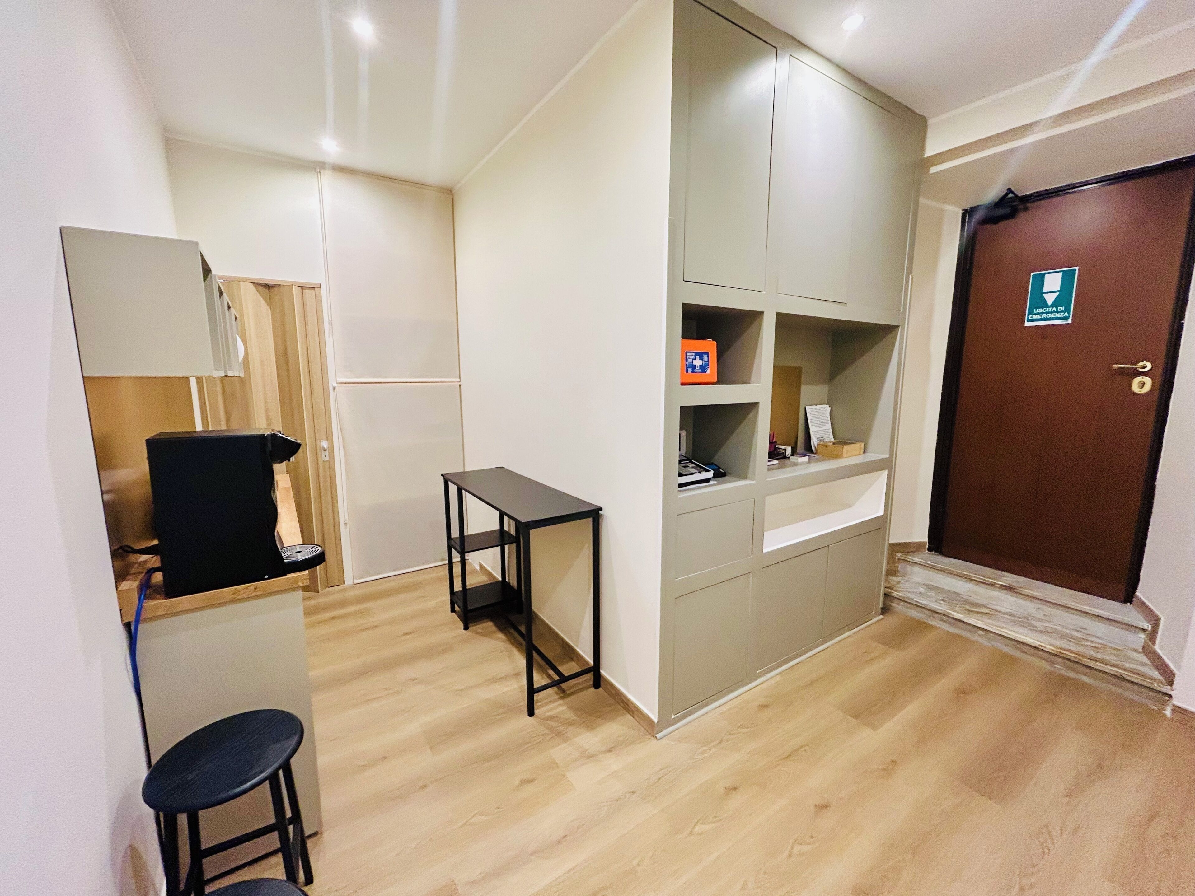 A small, compact room with a kitchenette, a single stool, a small table, and a door with an emergency exit sign.