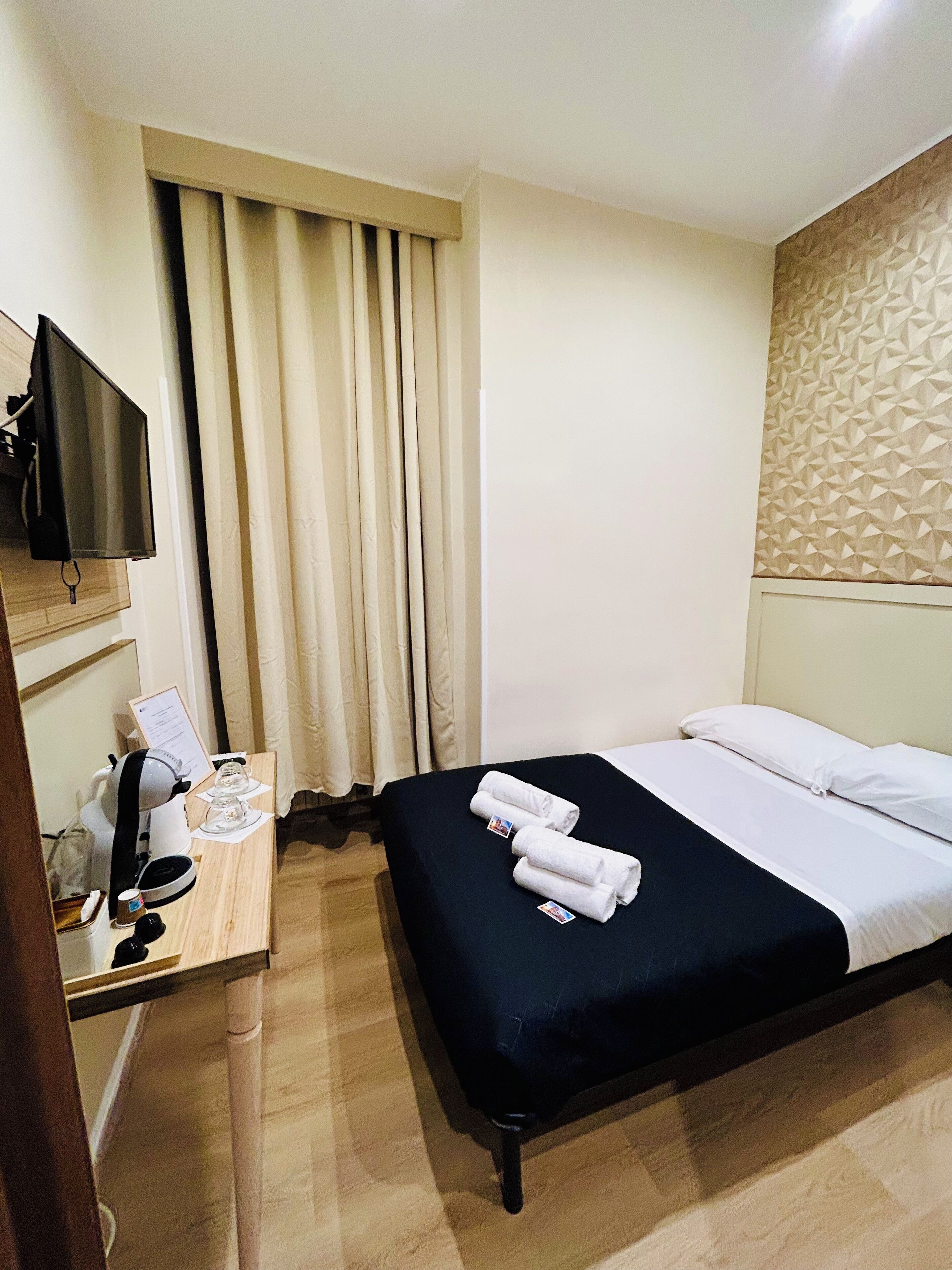 Habitación de hotel con cama, escritorio, teléfono, cafetera, televisión montada en la pared y ventana con cortinas.