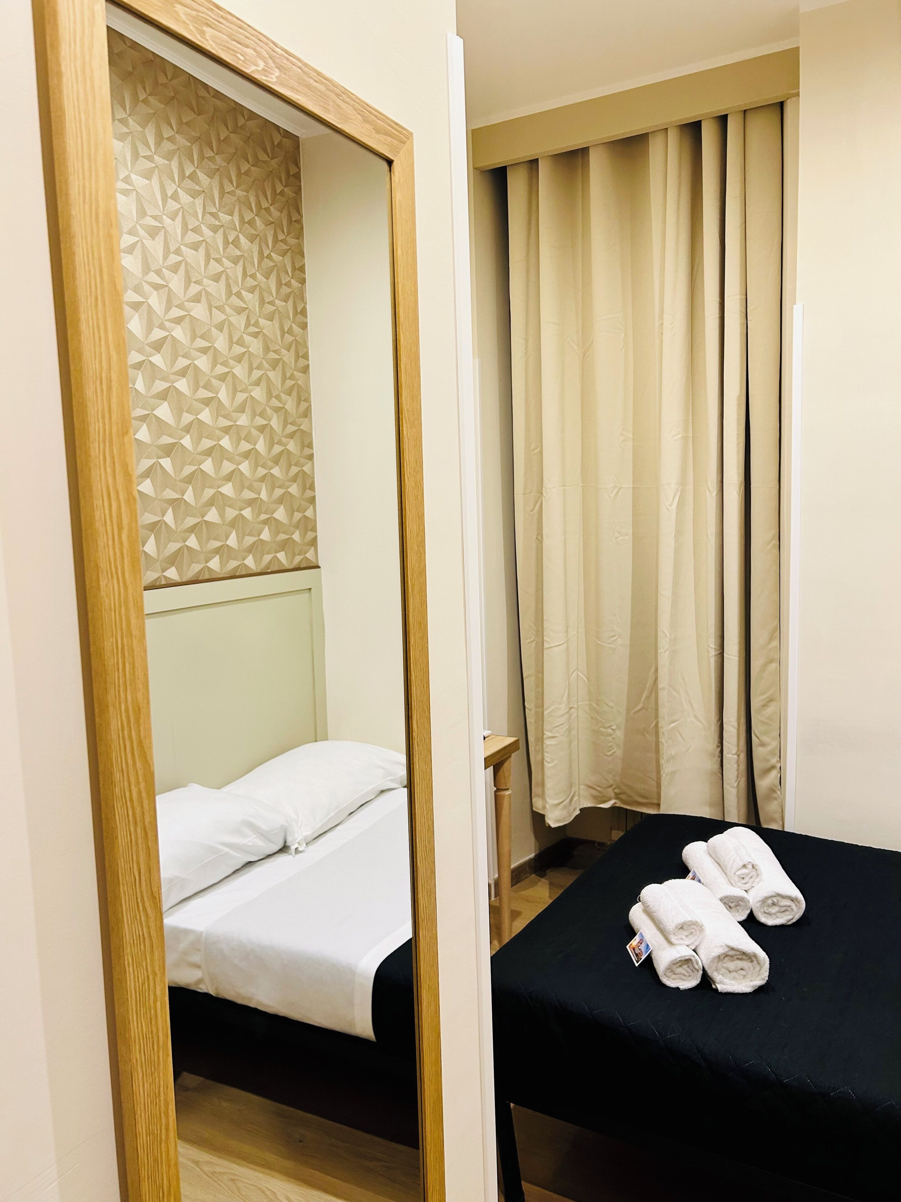 A hotel room with a bed, a small table with towels, a mirror, and a window with curtains.