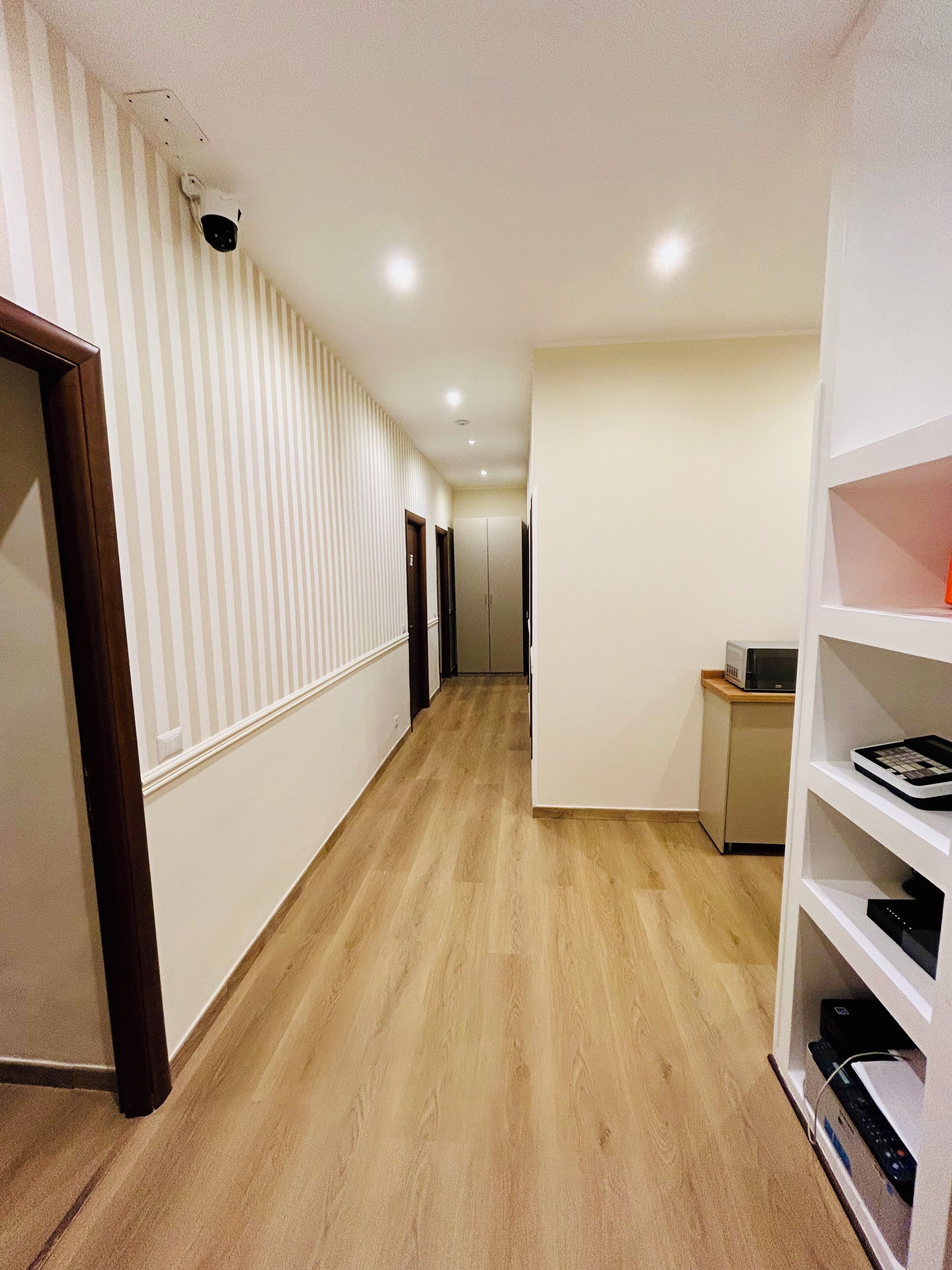 A hallway with wooden flooring, white walls, and a white shelf unit with electronic devices.