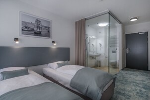 Quarto de hotel com duas camas, área de chuveiro com paredes de vidro, um quadro na parede e uma porta escura.