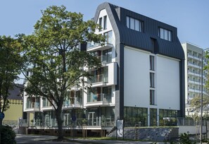 Un immeuble moderne à plusieurs étages, doté de balcons, de grandes fenêtres et d’un arbre devant.