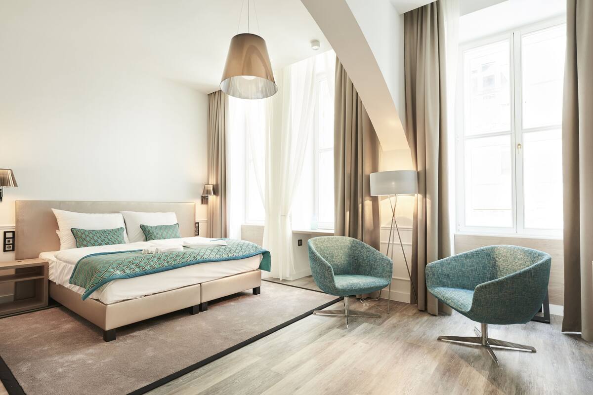 A modern bedroom with a bed, two chairs, a floor lamp, and large windows with curtains.