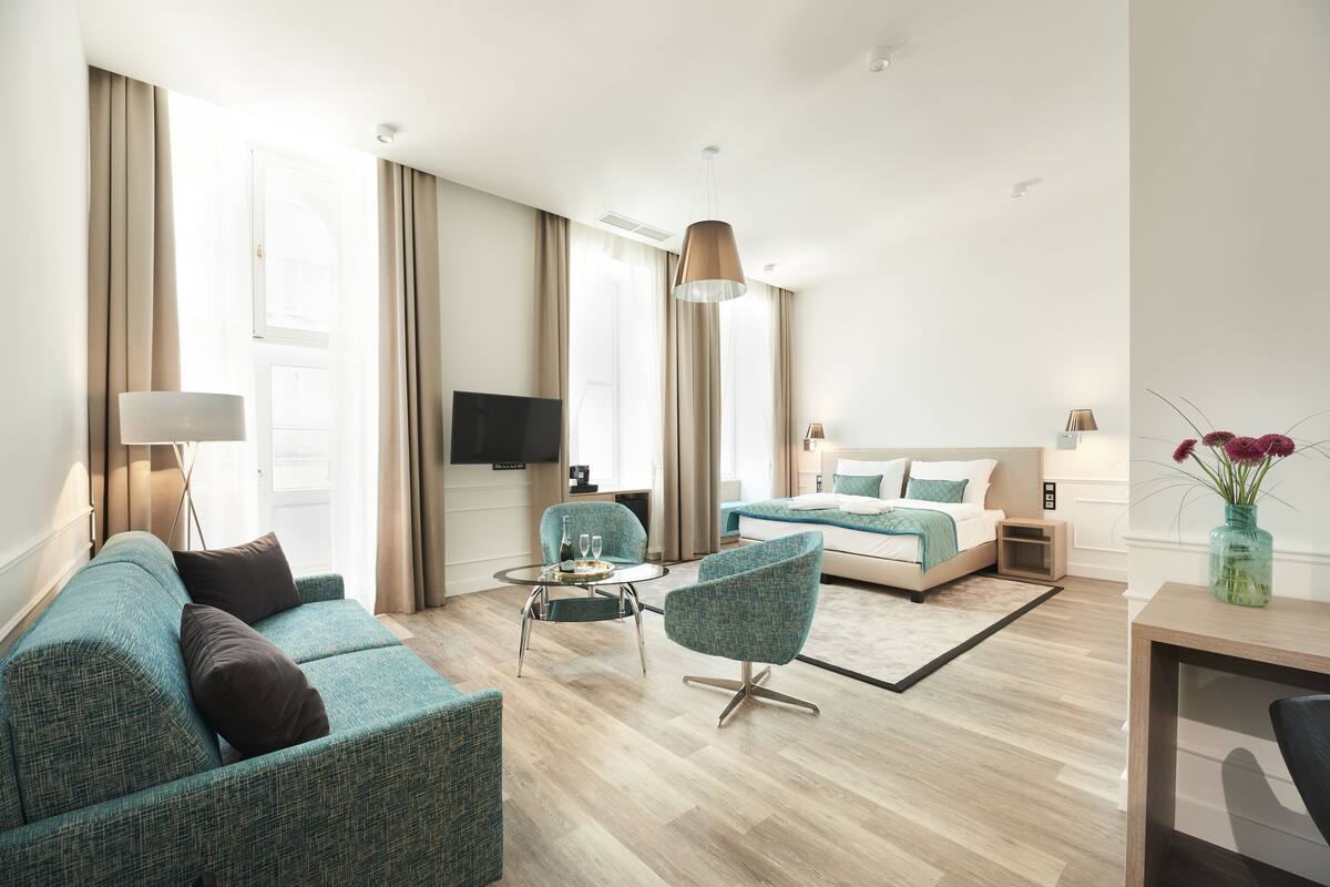 A modern hotel room with a sofa, armchair, bed, TV, and a dining area.