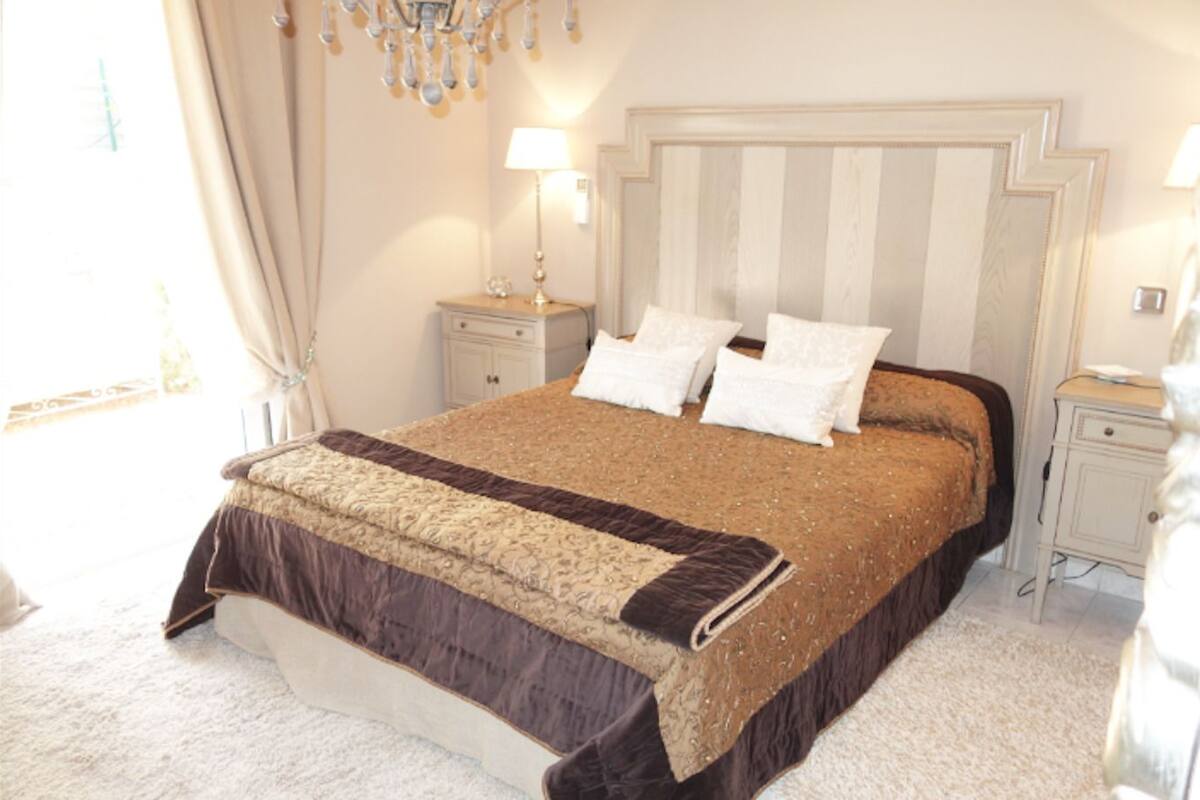 Ein ordentlich eingerichtetes Schlafzimmer mit einem großen Bett, einem Nachttisch, einer Stehlampe und einer Kristallleuchtre.