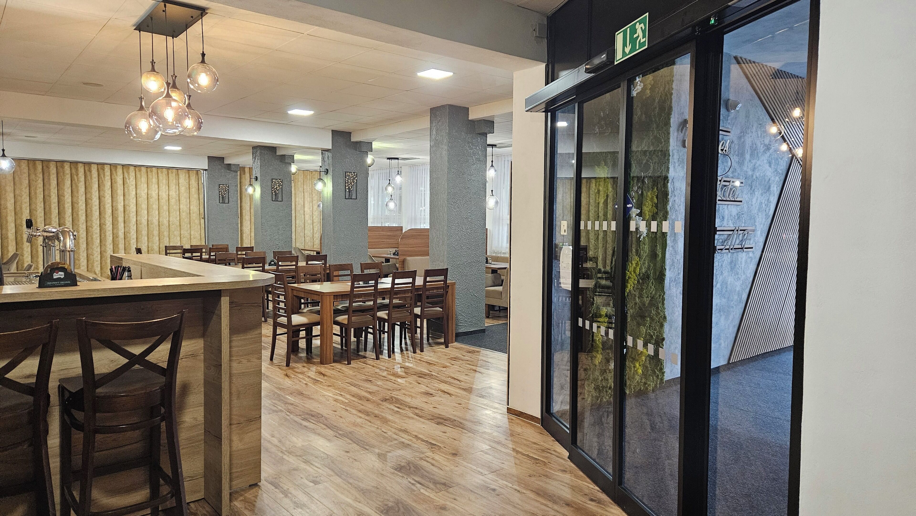 Un interior de restaurante moderno con mesas y sillas de madera, una zona de bar y un amplio ventanal con vistas a la vegetación.