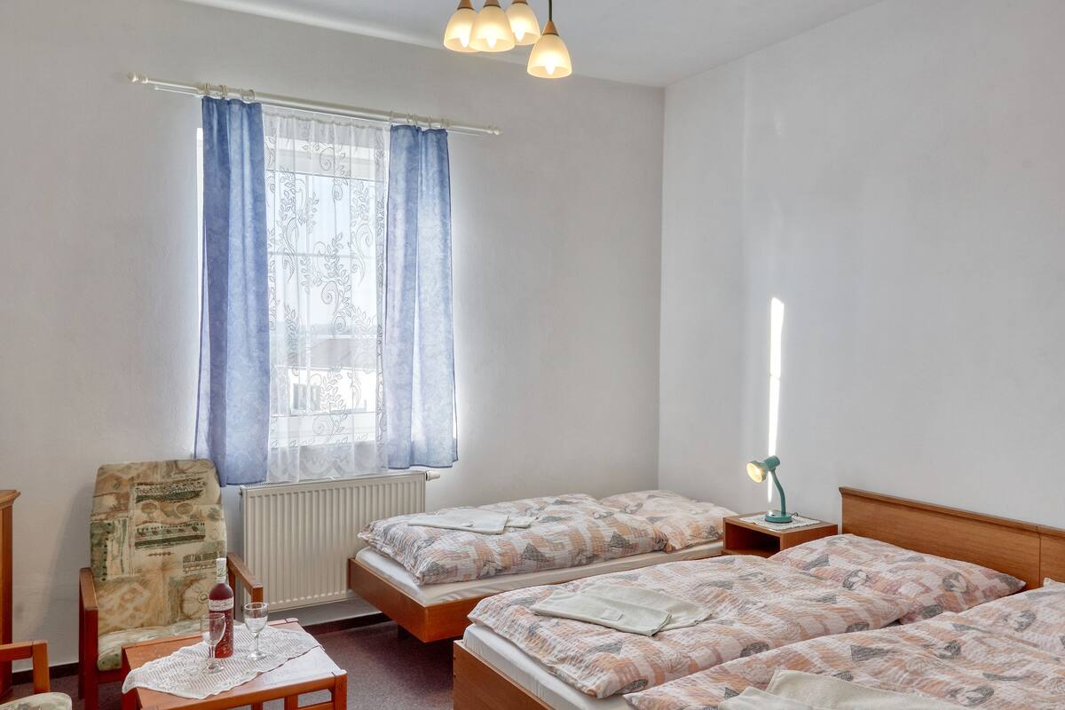 Una habitación con dos camas, una silla, una mesa con una botella y vasos, y un ventanal con cortinas.