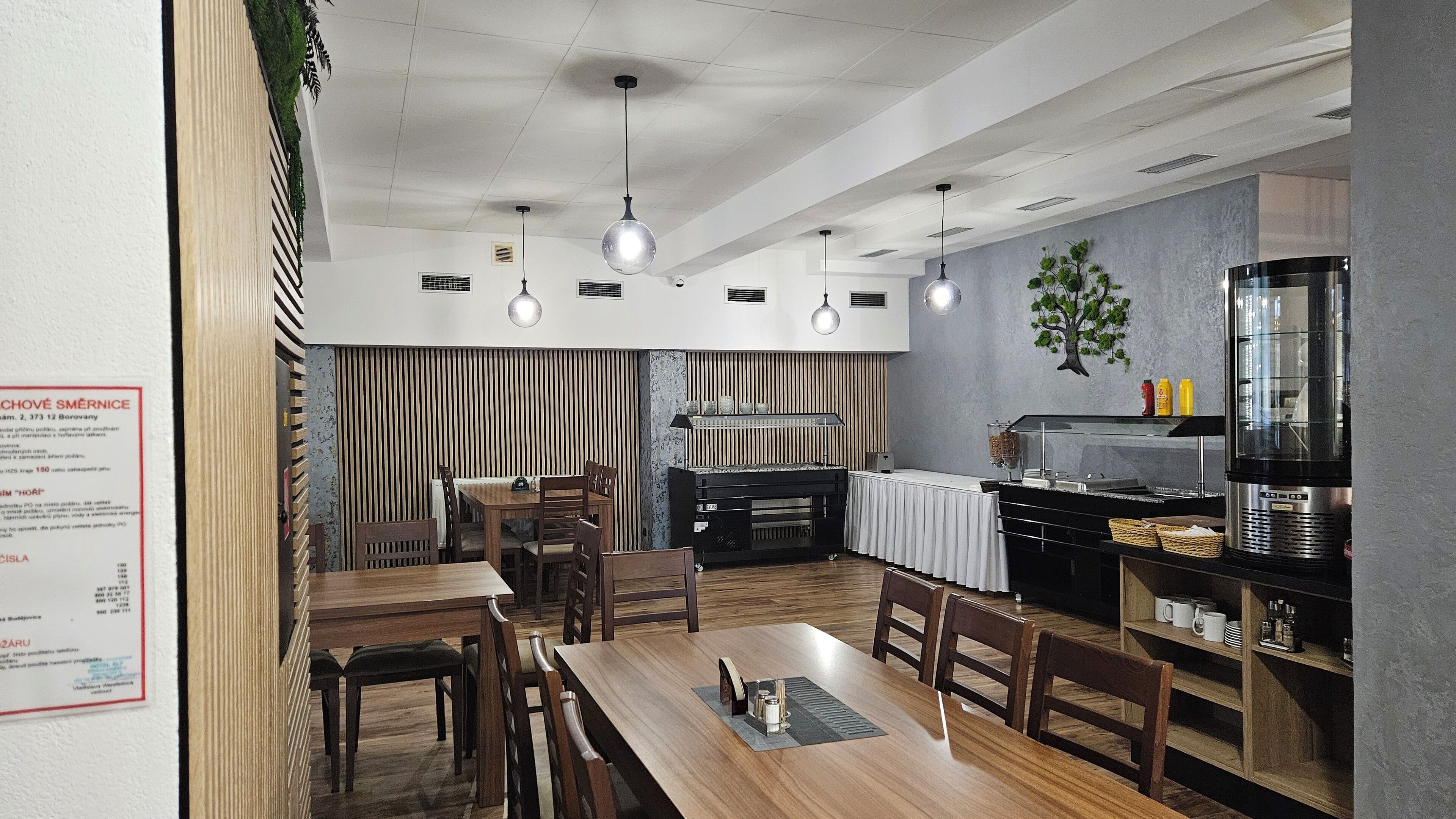 A restaurant interior with wooden tables and chairs, hanging pendant lights, a buffet counter, and a refrigerator.