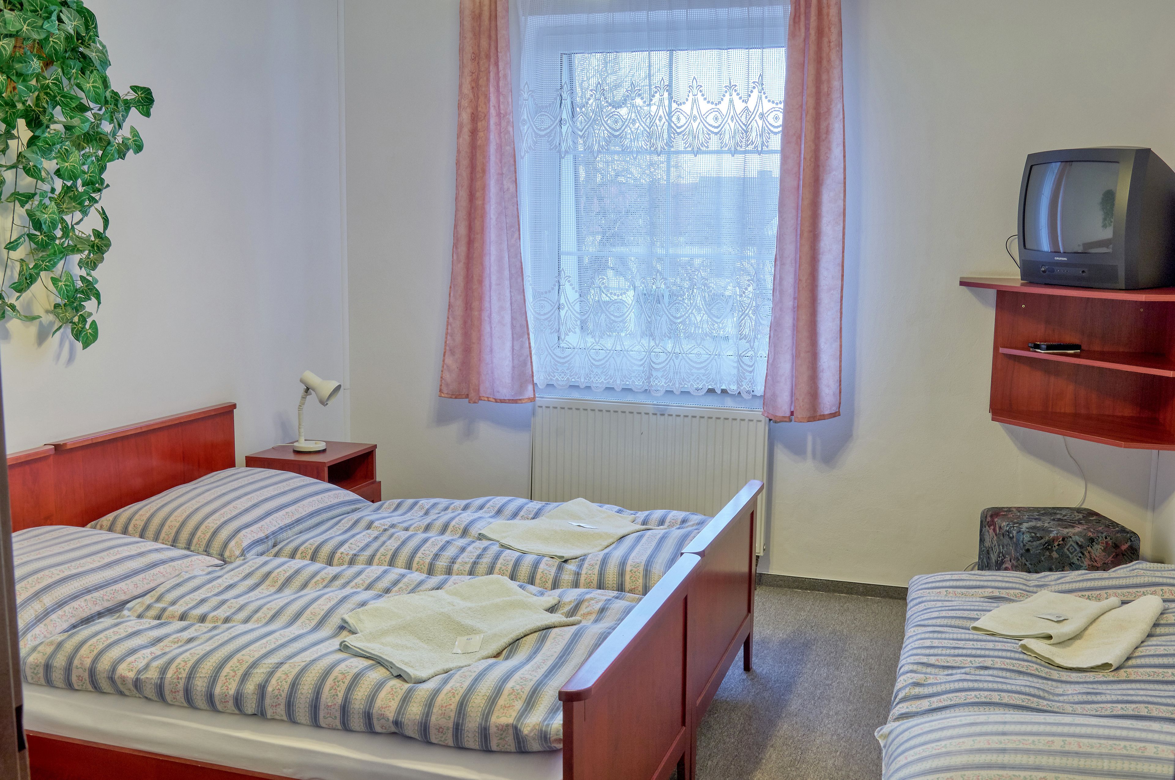Una habitación con dos camas, un televisor en un estante, un radiador y una ventana con cortinas.