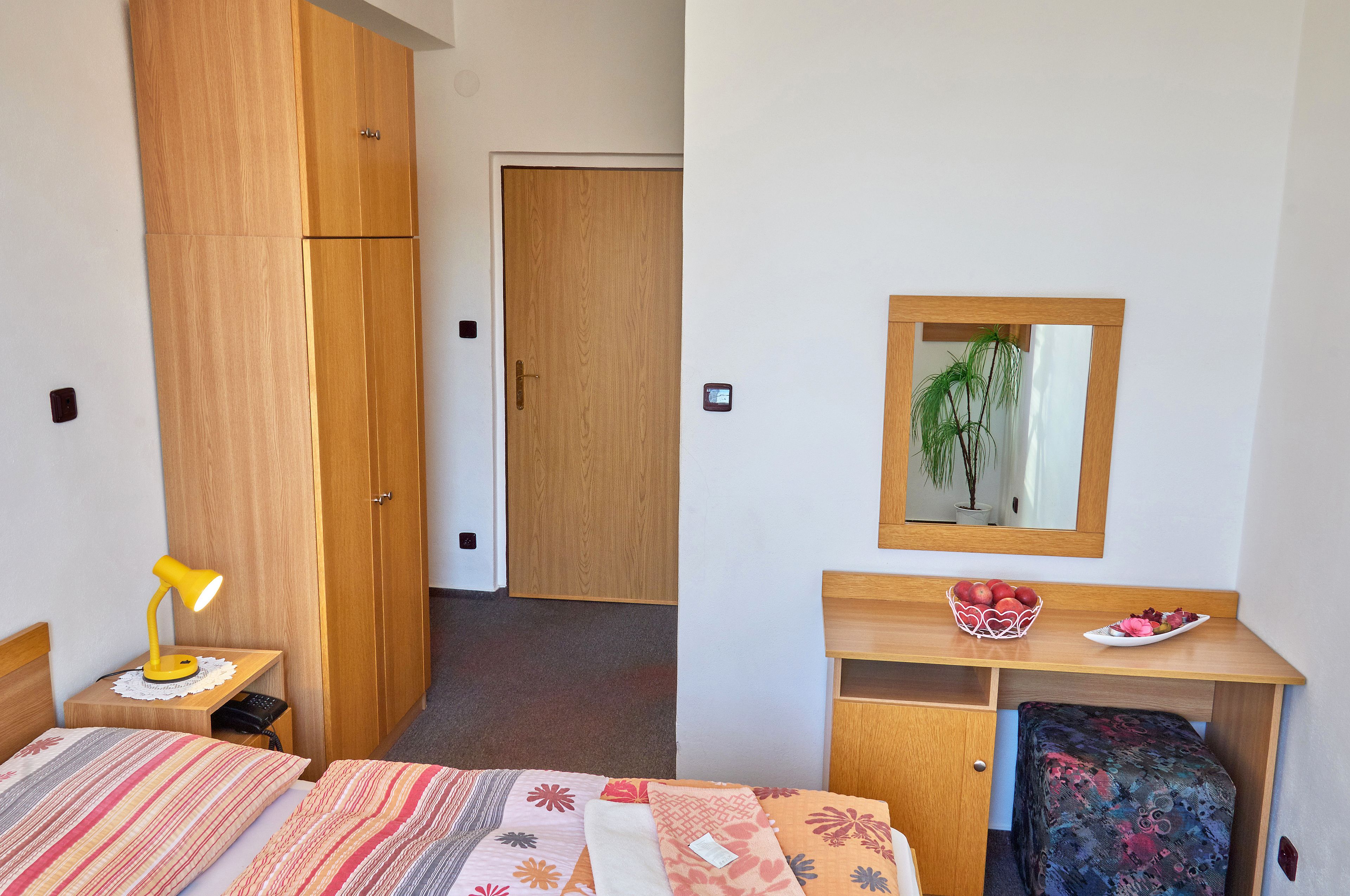 Ein Hotelzimmer mit Bett, Schreibtisch, Kleiderschrank, Nachttischlampe, Spiegel und einer kleinen Pflanze.