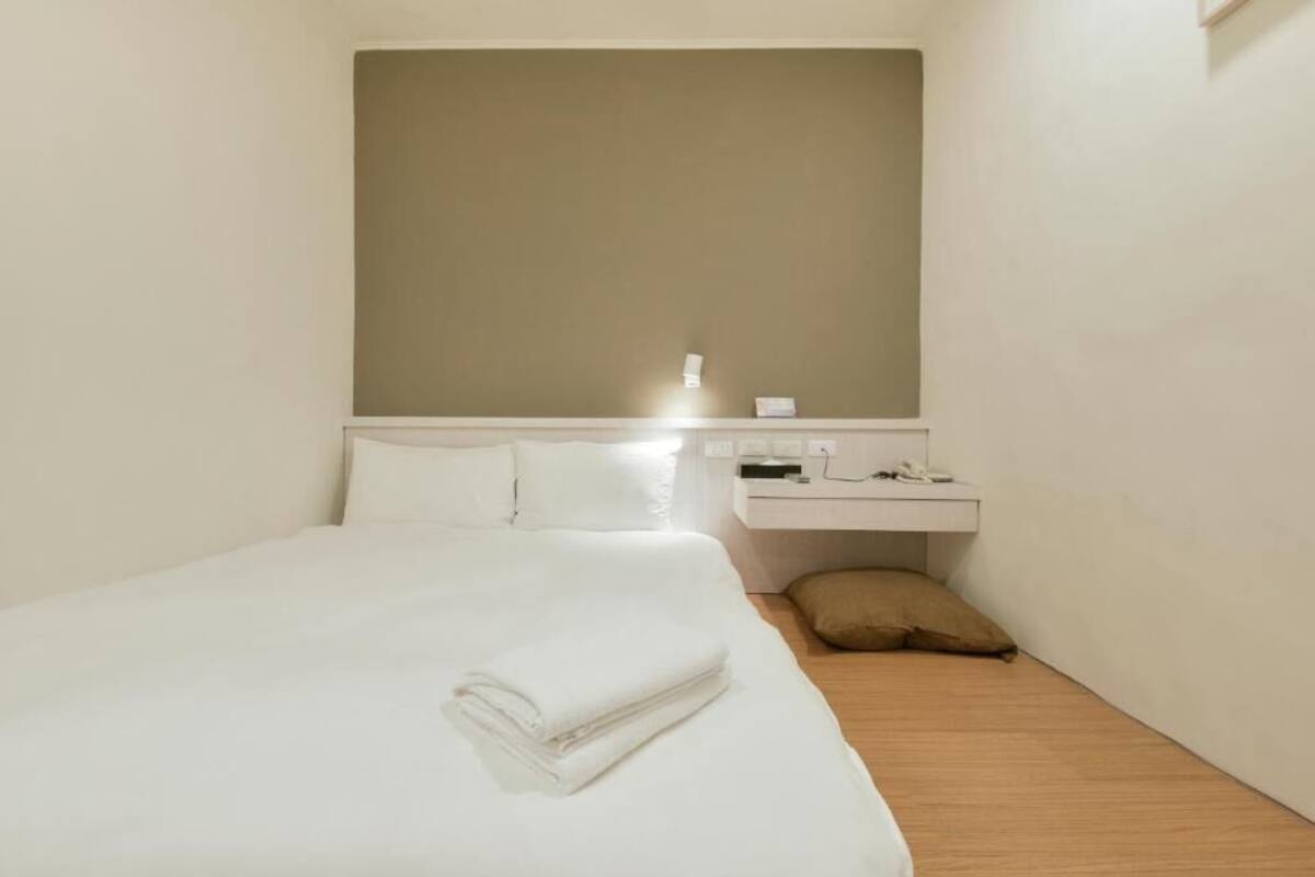 A hotel room with a neatly made bed, a bedside table with a lamp, and a small bedside shelf.