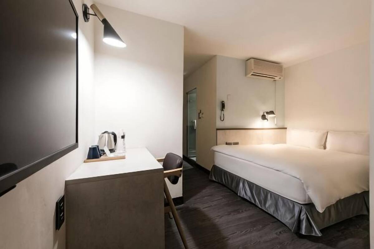 A hotel room with a bed, a desk with a lamp, a mirror, and a shower area.