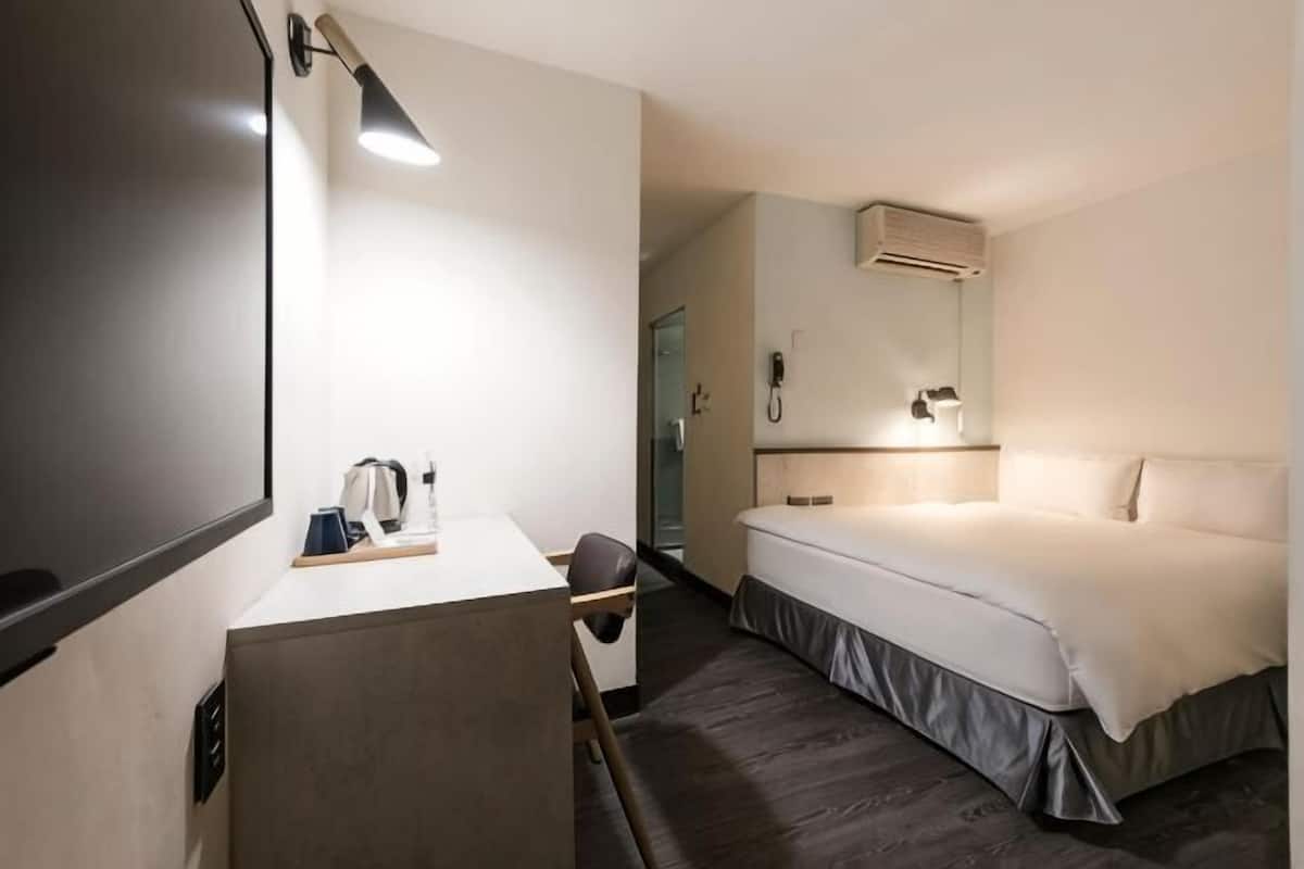 A hotel room with a bed, a desk with a lamp, a mirror, and a shower area.