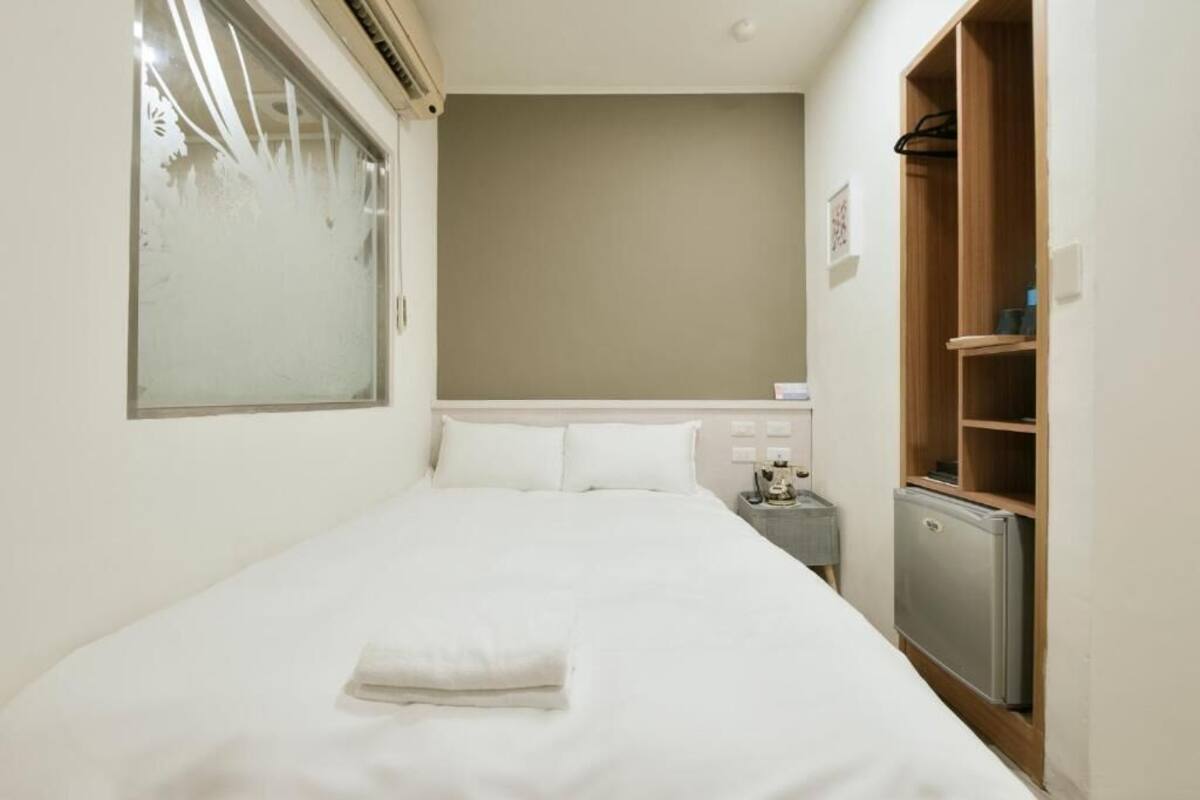 A small, neatly made room with a single bed, a nightstand, a small refrigerator, and a wooden wardrobe.