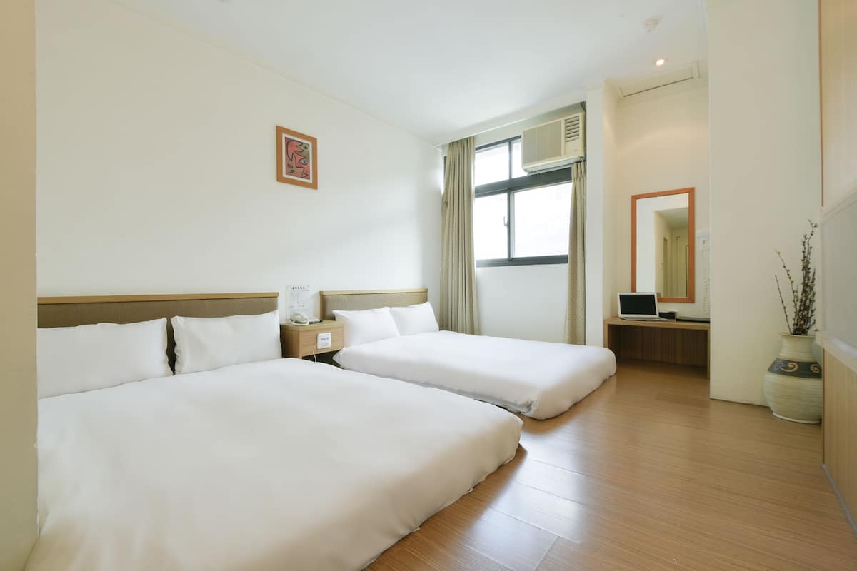 A hotel room with two beds, a desk with a laptop, a window with curtains, and a vase with branches.