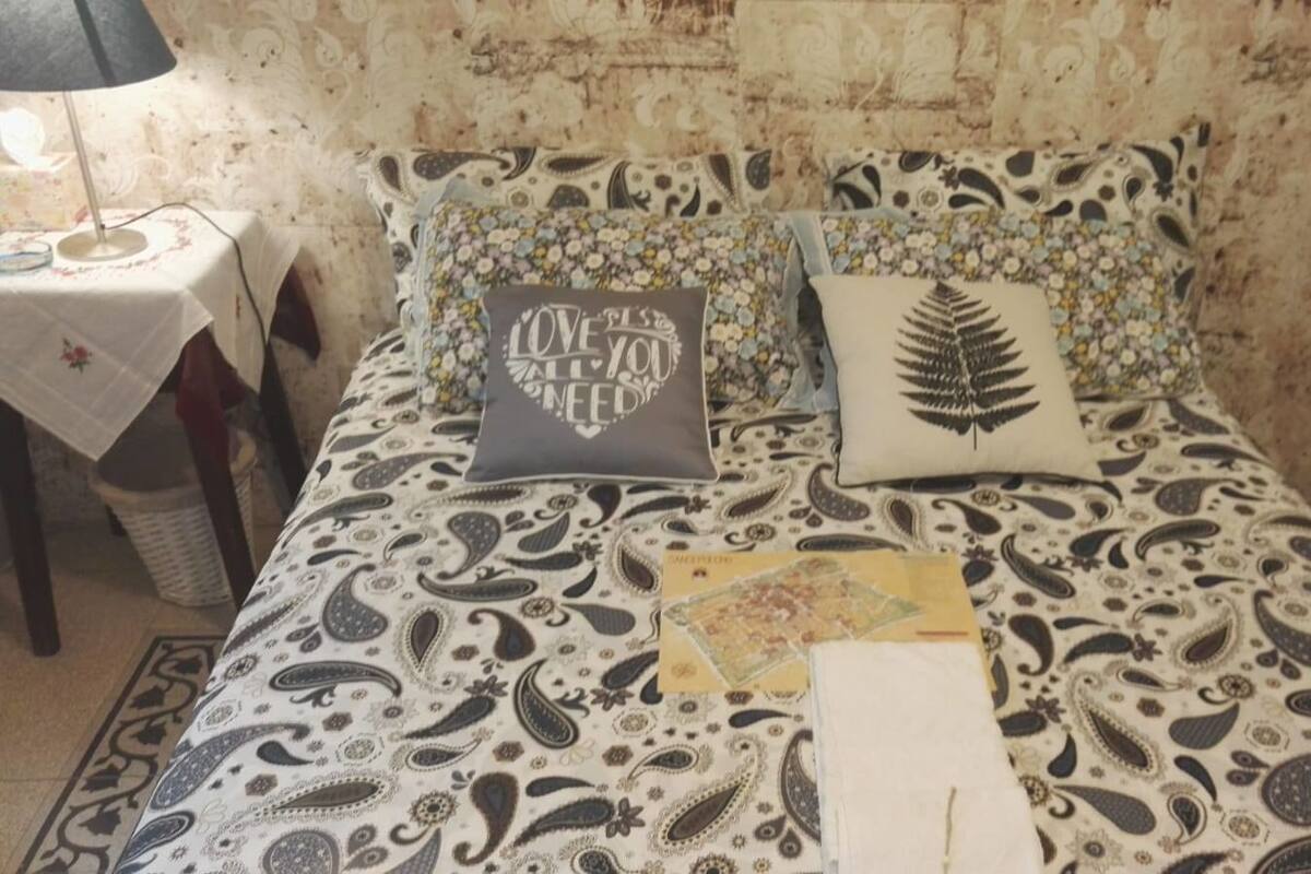 A bed with a patterned bedspread, two decorative pillows, a small table with a lamp, and a trash bin.