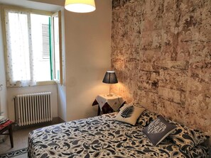 A bedroom with a patterned bedspread, a bedside lamp, a radiator, and a window with curtains. - Camere Mazzini 49 (Sansepolcro)