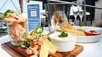 Una bandeja de mariscos con camarones, limón y ensalada sobre una tabla de madera, acompañada de un bol de salsa y pan, con un cartel de menú con código QR al fondo.
