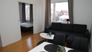 A modern living room with a black sofa, a small white table, and a glass coffee table. A bed is visible in the background, and the room has large windows with curtains. - Fastliving Apartment Hotel (Vasteras)