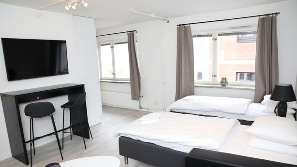 A modern hotel room with a flat-screen TV, two beds, a desk with chairs, and a window with curtains. - Fastliving Apartment Hotel (Vasteras)