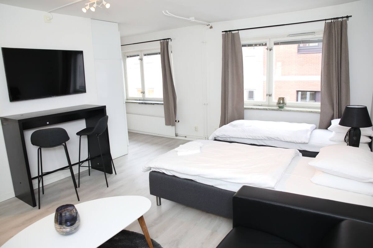 A modern hotel room with a flat-screen TV, two beds, a desk with chairs, and a window with curtains.