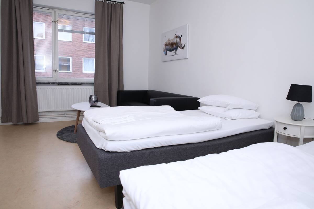 A hotel room with a bed, a small table, a chair, a lamp, and a window with curtains.