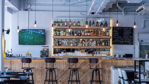 A bar area with multiple shelves of bottles, a television displaying a sports game, and a chalkboard menu listing cocktails.