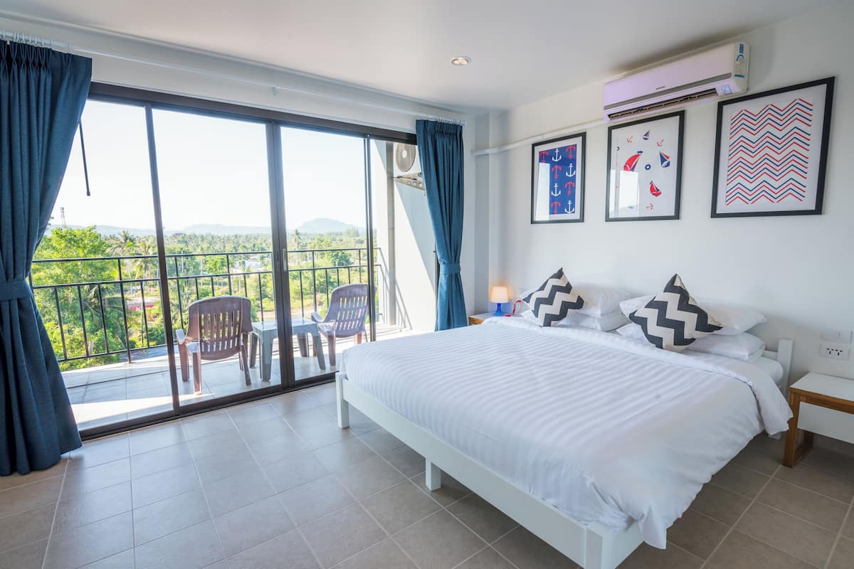 Une chambre avec un grand lit, un balcon avec des chaises et une vue sur la verdure.