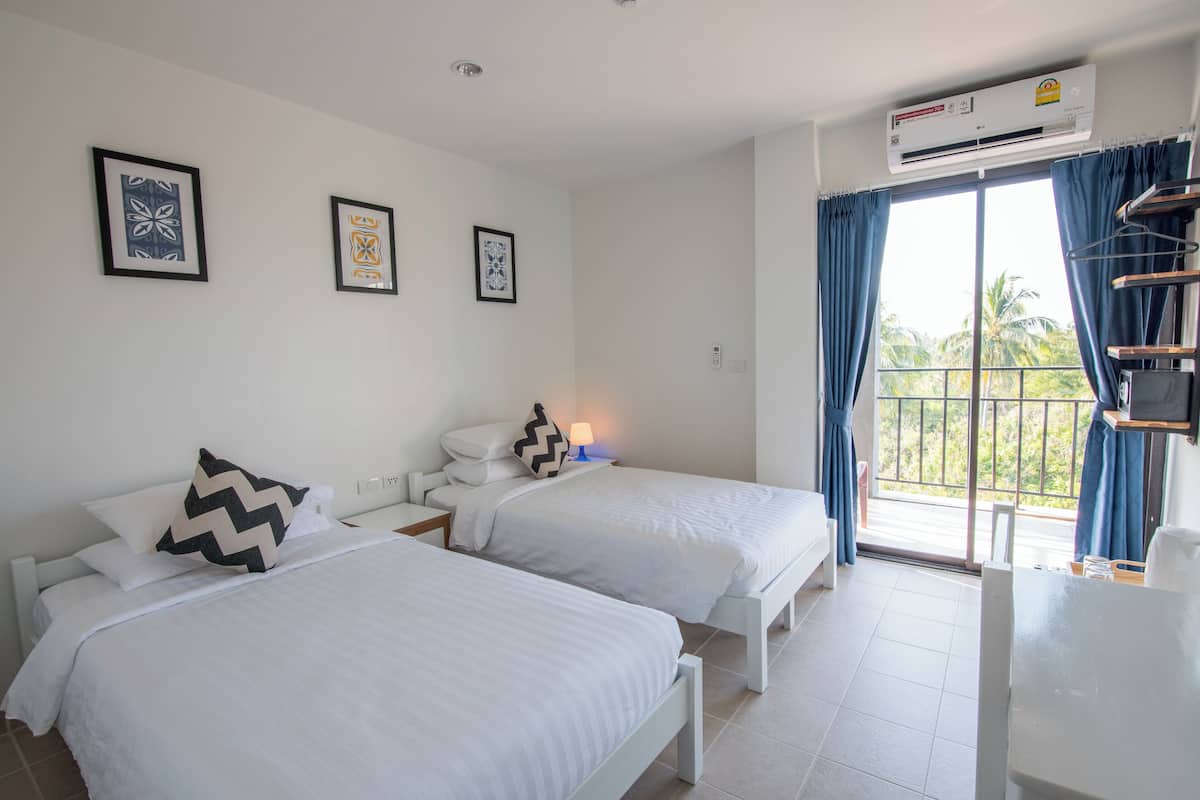 Une chambre d’hôtel avec deux lits, une fenêtre avec des rideaux, un appareil de climatisation et des œuvres d’art encadrées accrochées au mur.